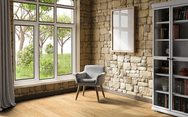 A cozy room with stone walls, a large window showing green trees outside, a gray armchair on a wooden floor, a white-framed bookshelf with books, and a blank picture frame on the wall.