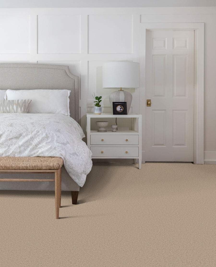 A bedroom with a white bed, wooden bench at the foot, nightstand with a lamp, small potted plant, and framed photo, against a white paneled wall and closed white door on beige carpet.