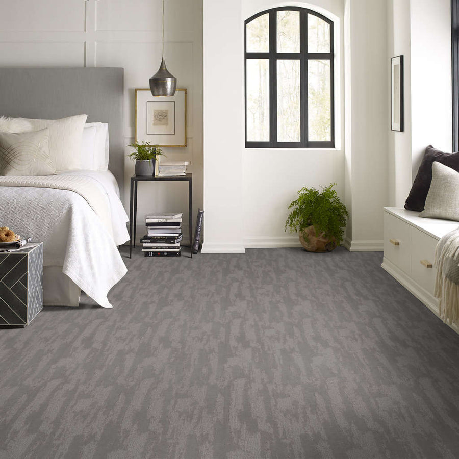 A modern bedroom with light walls, a bed with white bedding, a black nightstand with decor, a large window, a potted plant, and gray textured carpet flooring.