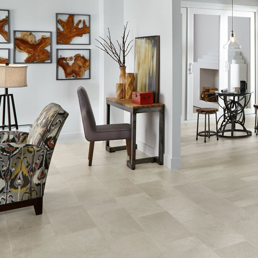 A modern living space with light beige tile flooring, a patterned armchair, a gray upholstered chair at a wooden desk, abstract wall art, and a glimpse of a dining area with stools and a fireplace.