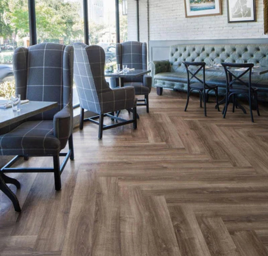 A modern café features checkered upholstered armchairs, black wooden tables, and a long green tufted bench against a white brick wall. Large windows highlight the wood-patterned herringbone Commercial Vinyl flooring.