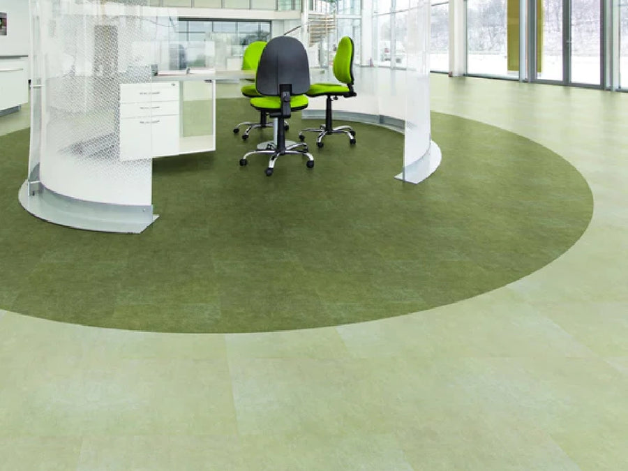 A modern office features Commercial Vinyl green flooring, glass partitions, and two green office chairs at a desk near large windows with abundant natural light.