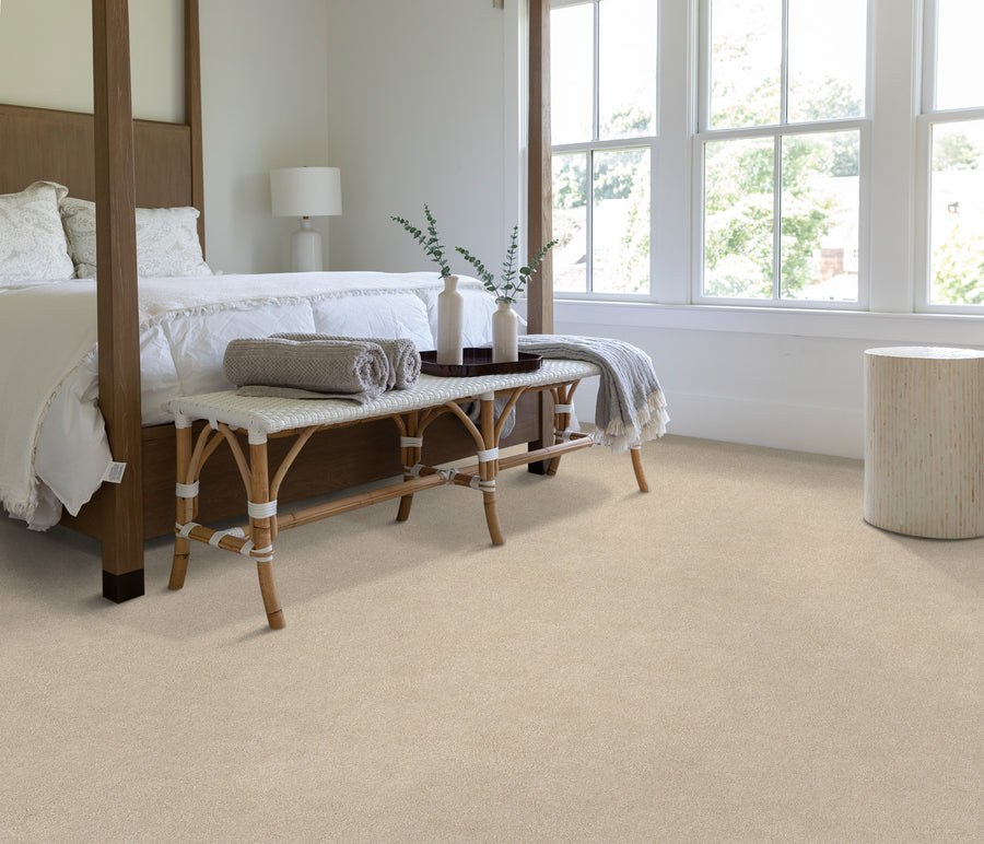 Bright bedroom with a four-poster bed, white bedding, a wooden bench with rolled towels at the foot, a round side table, and large windows letting in natural light onto a beige carpeted floor.