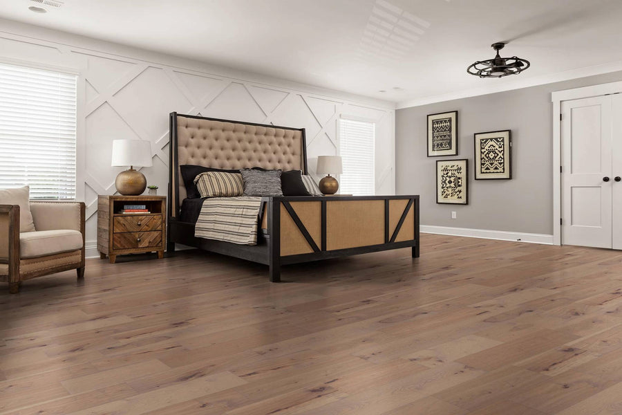 A modern bedroom with a large upholstered bed, beige armchair, two nightstands with lamps, wood floors, gray and white walls, framed art, and double white doors. Light streams in from two windows with blinds.