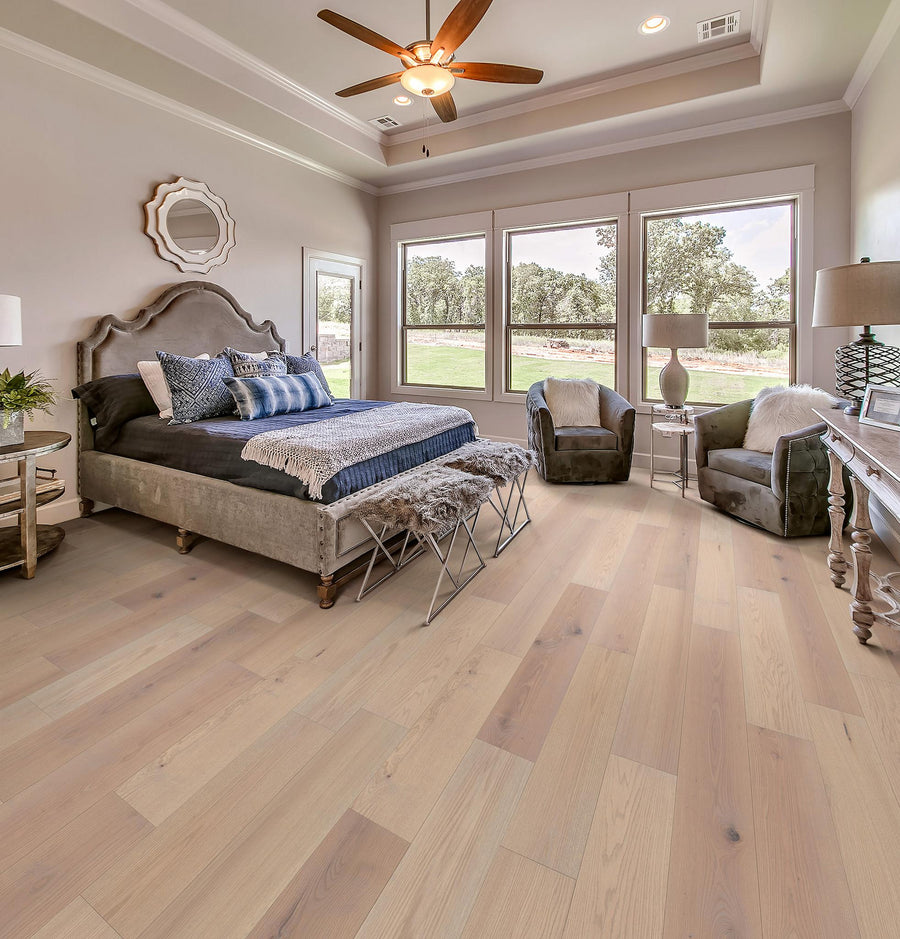 A spacious, elegant bedroom with a large upholstered bed, soft neutral bedding, two armchairs near tall windows, light wood floors, and a ceiling fan, all filled with natural light.