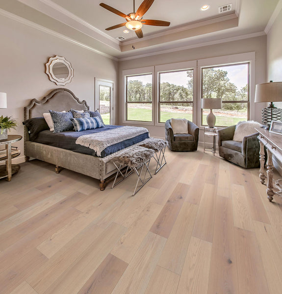 A spacious, elegant bedroom with a large upholstered bed, soft neutral bedding, two armchairs near tall windows, light wood floors, and a ceiling fan, all filled with natural light.
