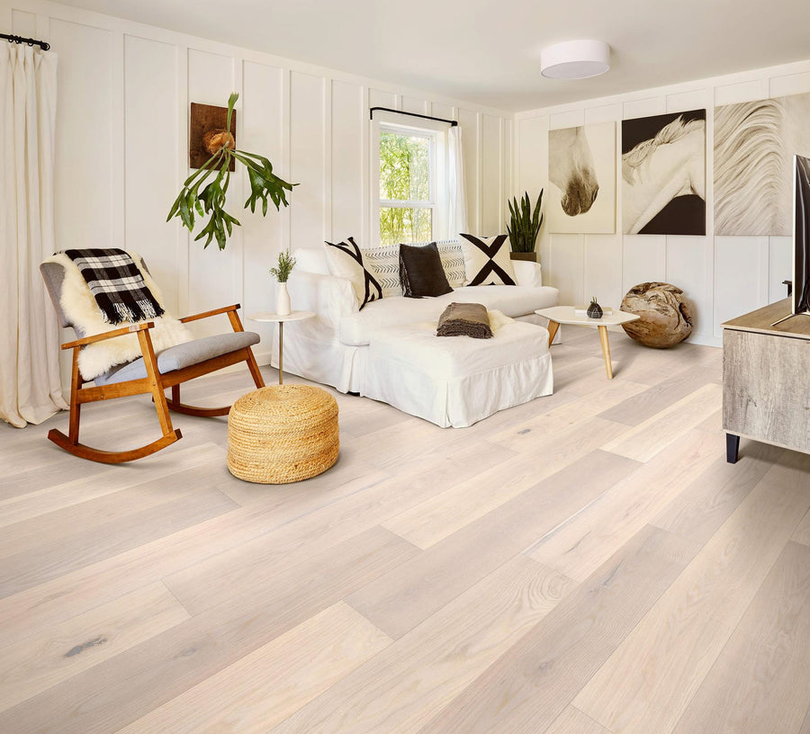 Bright, modern living room with light wood floors, a white sofa, a wooden rocking chair, potted plants, a woven ottoman, and large horse artwork on the wall. Sunlight streams in through a window.