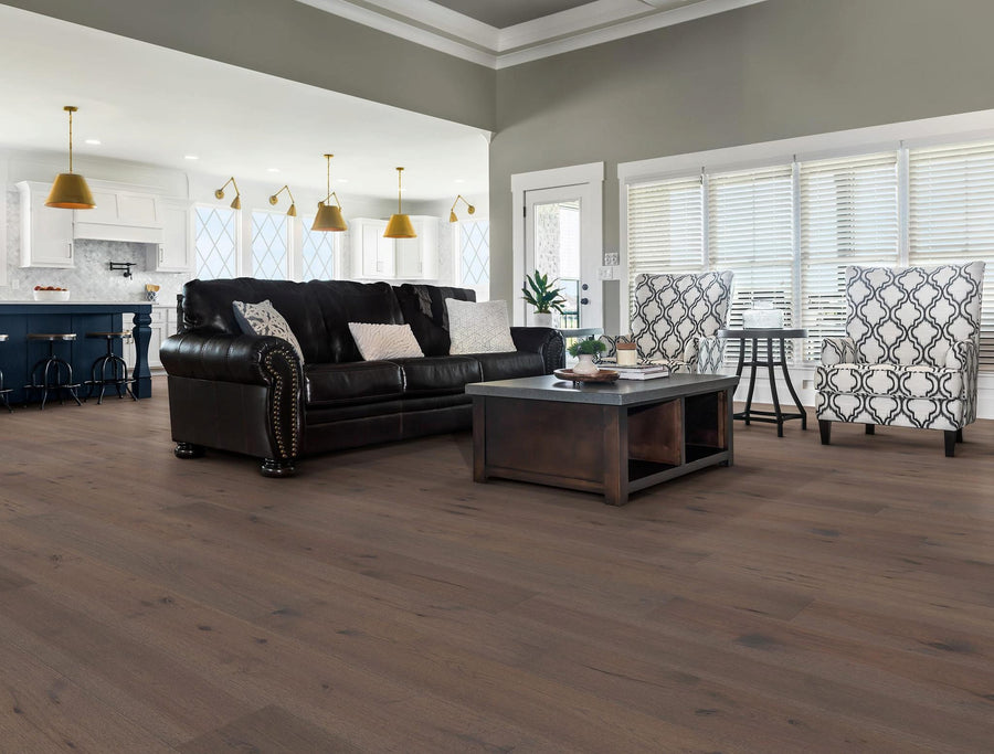 A spacious, modern living room with wood floors, a dark leather sofa, two patterned armchairs, a wooden coffee table, and large windows. An open kitchen with pendant lights is visible in the background.