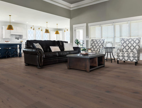 A spacious, modern living room with wood floors, a dark leather sofa, two patterned armchairs, a wooden coffee table, and large windows. An open kitchen with pendant lights is visible in the background.