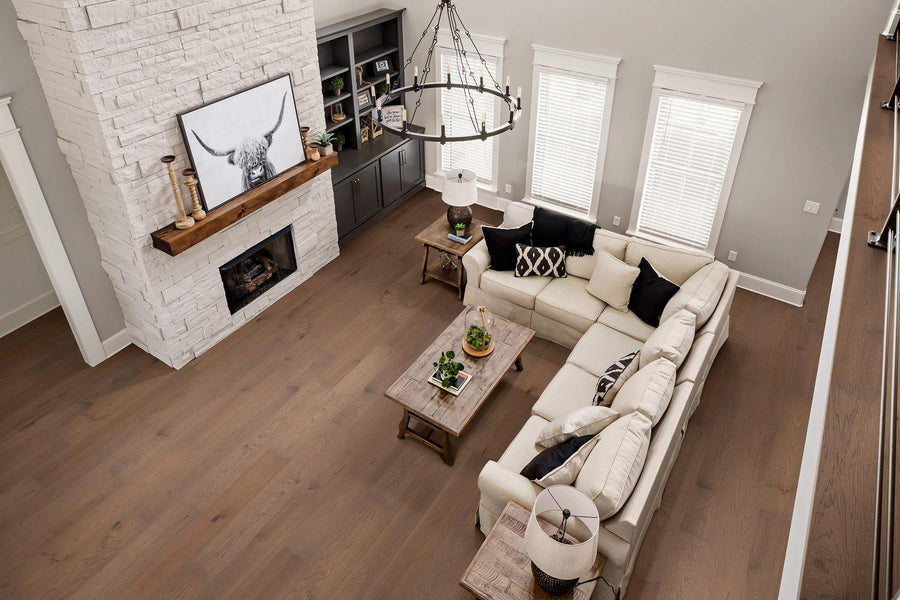 A modern living room with light wood floors, a white sectional sofa, a wooden coffee table, a white stone fireplace, built-in shelves, and large windows with white blinds. A black chandelier hangs above the seating area.