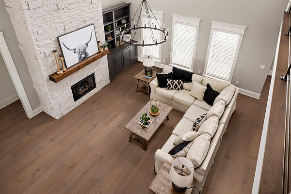 A modern living room with light wood floors, a white sectional sofa, a wooden coffee table, a white stone fireplace, built-in shelves, and large windows with white blinds. A black chandelier hangs above the seating area.