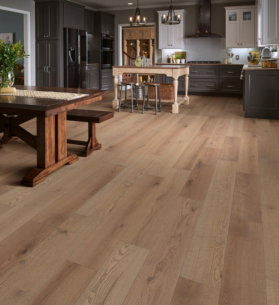 A spacious kitchen and dining area with wide light brown wooden flooring, dark cabinetry, a farmhouse table, modern kitchen island, pendant lights, and various kitchen appliances and decor.