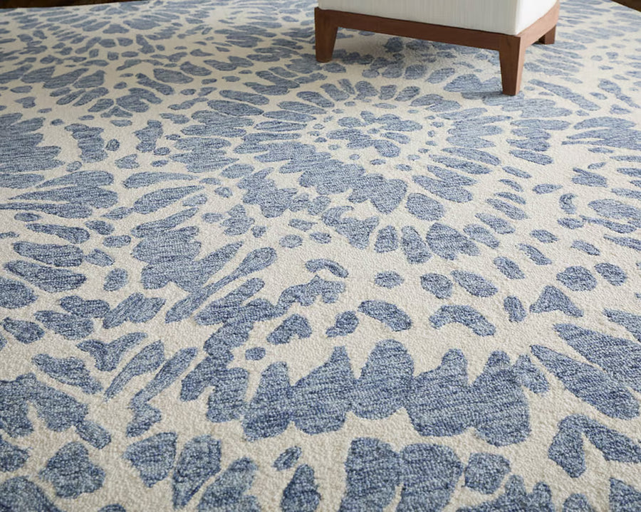 A close-up of a rug with a blue and white abstract, swirl-like pattern. The corner of a wooden chair with a white cushion is visible in the upper right corner.