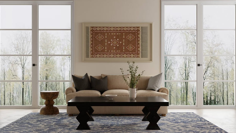 A modern living room with a beige sofa, patterned pillows, a dark geometric coffee table, a small side table, a vase with branches, a blue patterned rug, and large windows showing trees outside. A framed textile hangs on the wall.