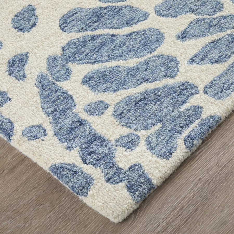 Close-up of a cream-colored area rug with irregular blue spots and a textured, looped surface, placed on a light brown wooden floor.