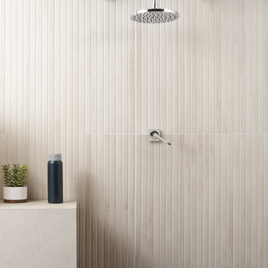 A modern shower with a round overhead showerhead, a single handle faucet on a beige textured tile wall, and a small ledge holding a potted plant and a black bottle.