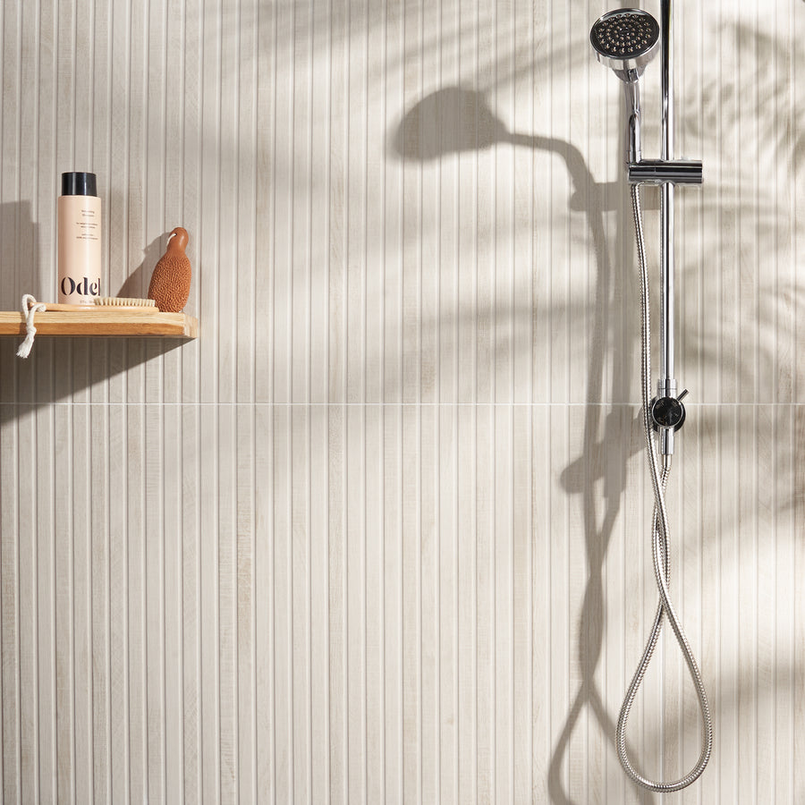 A modern shower with a chrome showerhead and hose against a textured beige tiled wall; a wooden shelf holds a bottle and a small decorative bird. Sunlight casts soft shadows on the wall.