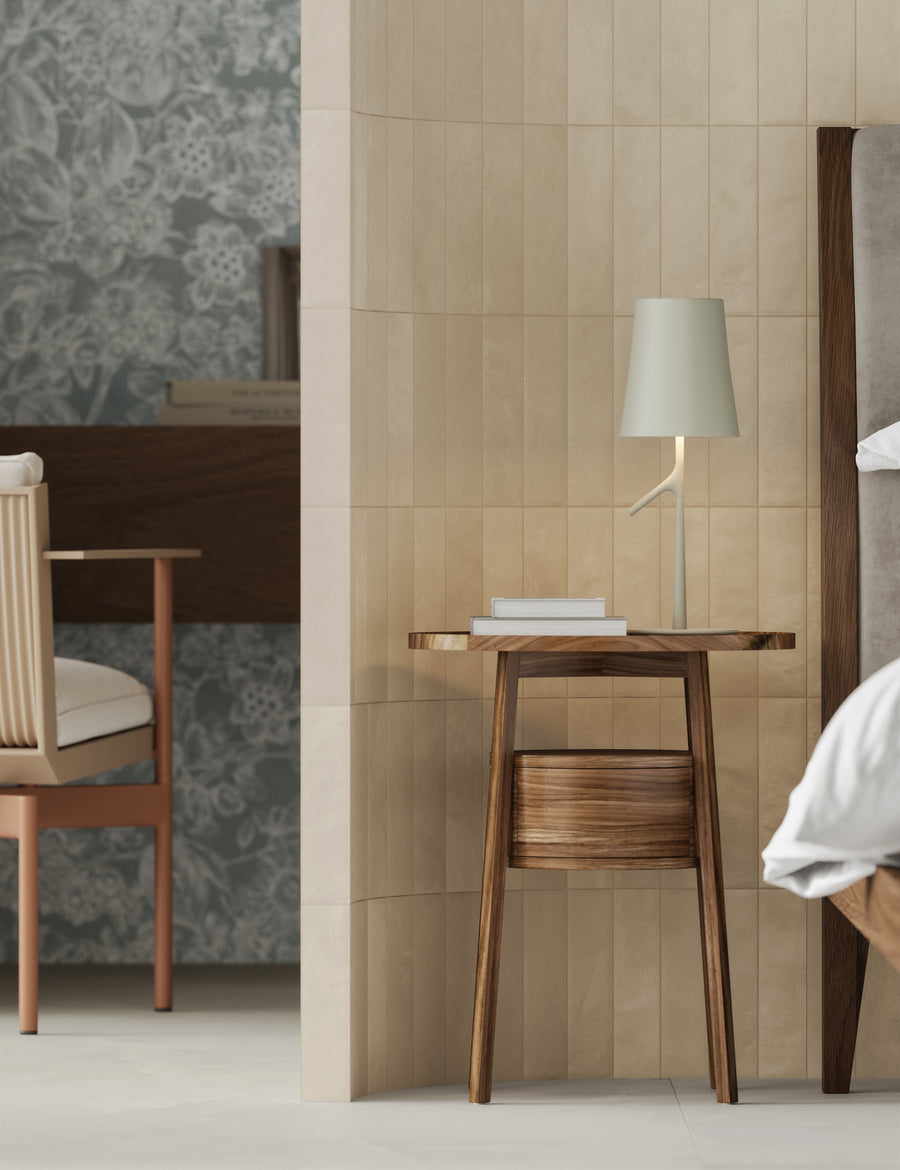 A modern bedroom scene with a wooden nightstand holding a white book and a minimalist lamp, next to a bed and a wall divider. In the background, theres a desk, chair, and floral wallpaper.