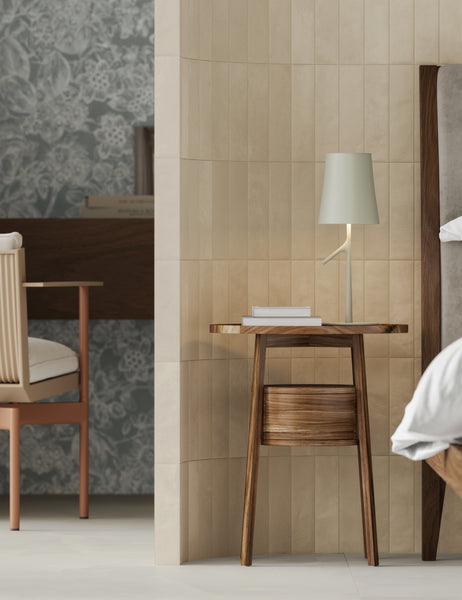A modern bedroom scene with a wooden nightstand holding a white book and a minimalist lamp, next to a bed and a wall divider. In the background, theres a desk, chair, and floral wallpaper.
