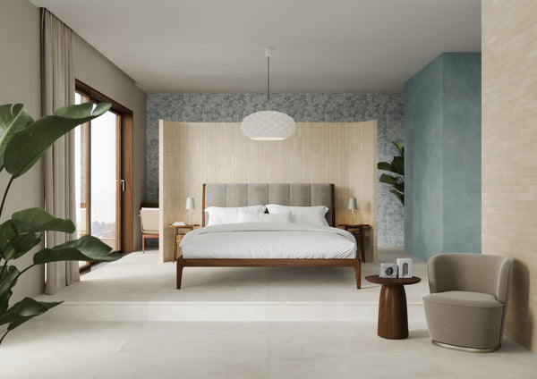 A modern bedroom with a wooden bed, white bedding, side tables, and a pendant light. There are large plants, a small round table with a camera, an armchair, and textured walls in beige and blue tones.