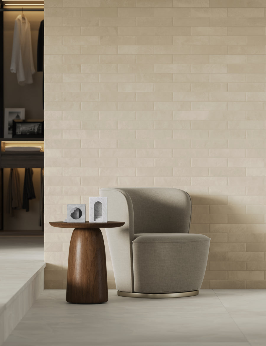 A modern beige armchair sits next to a round wooden side table with two decorative objects, against a light brick wall. An open closet with hanging clothes is visible in the background.