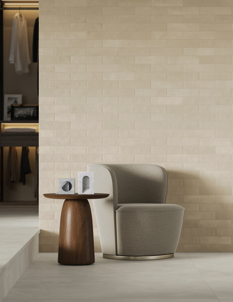 A modern beige armchair sits next to a round wooden side table with two decorative objects, against a light brick wall. An open closet with hanging clothes is visible in the background.