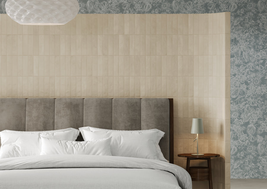 A modern bedroom with a gray upholstered headboard, white bedding, a small round wooden side table with a pale green lamp, and textured beige and blue floral wallpaper.