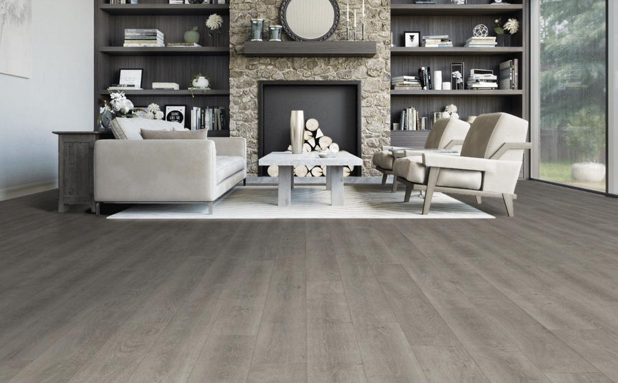 A modern living room with light gray wood floors, a stone fireplace, a beige sofa, two matching armchairs, a white coffee table, and built-in bookshelves filled with decor and books. Large windows let in natural light.