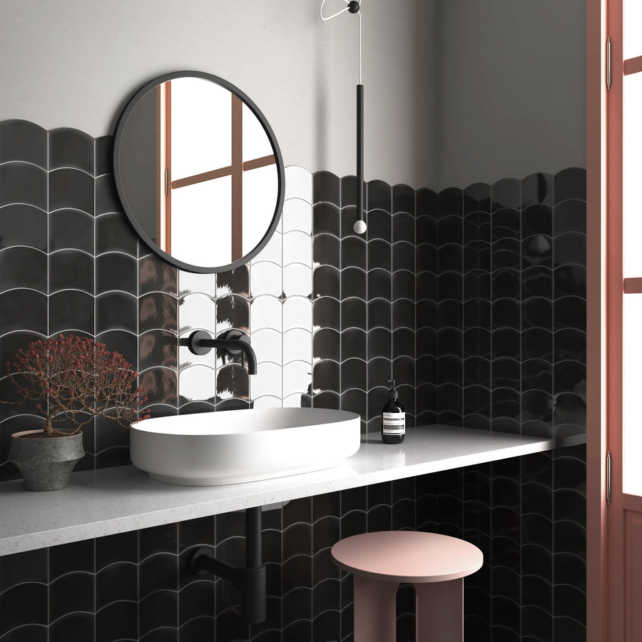 A modern bathroom with black scalloped tile walls, a round mirror, a white vessel sink, matte black faucet, potted plant, soap dispenser, and a pink stool next to a window with natural light.