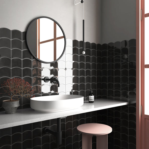 A modern bathroom with black scalloped tile walls, a round mirror, a white vessel sink, matte black faucet, potted plant, soap dispenser, and a pink stool next to a window with natural light.