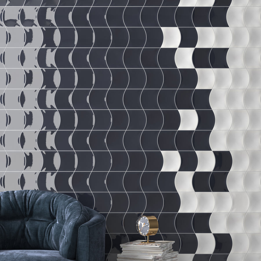 A modern wall with wavy, glossy tiles in shades of black, gray, and white. In front sits a dark blue tufted chair, a small stack of books, and a golden clock on a table.