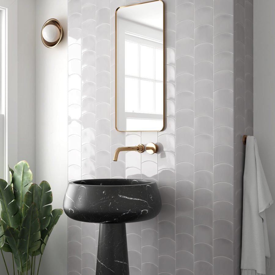 Modern bathroom with glossy white scalloped tiles, a black marble pedestal sink, gold fixtures, a rectangular mirror, a wall sconce, a white towel on a hook, and a green potted plant in the corner.