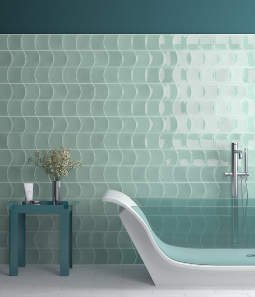Modern bathroom with light teal wavy-patterned wall tiles, a sleek white bathtub filled with water, a teal side table holding a vase with branches and a bottle, and a silver floor-mounted tub faucet.