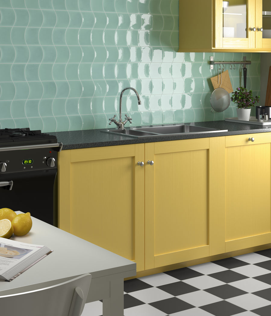 A kitchen with yellow cabinets, black countertop, stainless steel sink, mint green wavy tile backsplash, checkered black-and-white floor, and lemons on a white table. Some kitchen utensils and a small plant are visible.