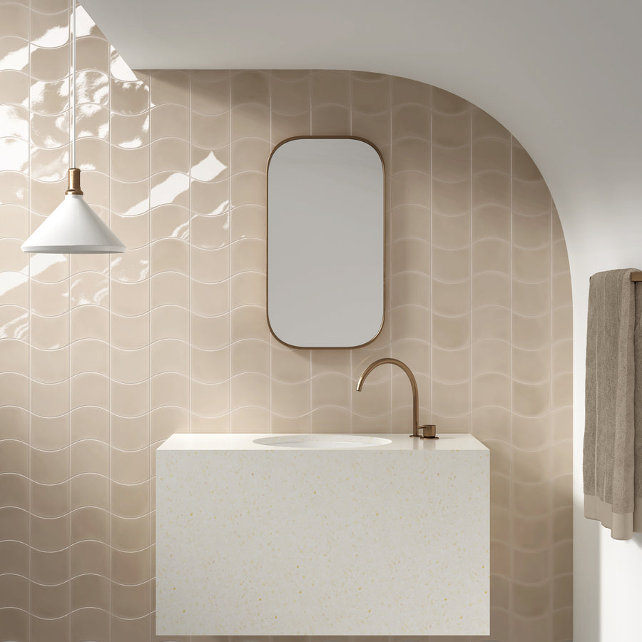 Modern bathroom with glossy beige wavy tiles, a white rectangular sink with a gold faucet, a wall-mounted oval mirror, a white pendant light, and a beige towel on a rack. The design is minimal and elegant.