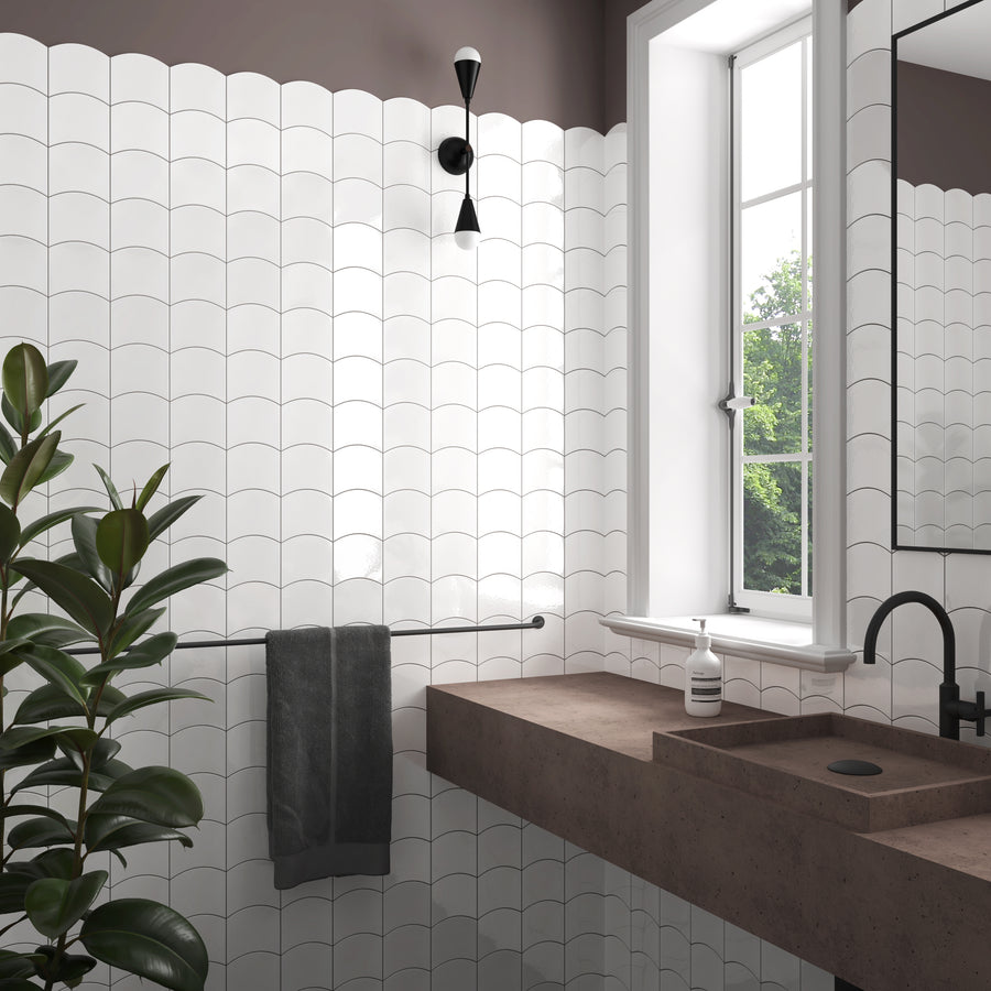 Modern bathroom with white scallop-patterned tiles, brown countertop with integrated sink, black faucet, wall-mounted towel bar with a dark towel, a plant, a window with greenery outside, and a wall sconce above.