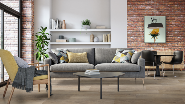 Modern living room with a gray sofa, patterned pillows, round coffee table, yellow armchair, large plant, wall shelves with books, exposed brick walls, and a dining area with black chairs and a round table.