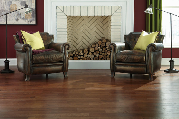 Two brown leather armchairs with yellow pillows face forward, positioned on Deepwood Trail 5″ Hickory Oak Hardwood flooring in front of a fireplace filled with stacked logs. Floor lamps flank each chair.