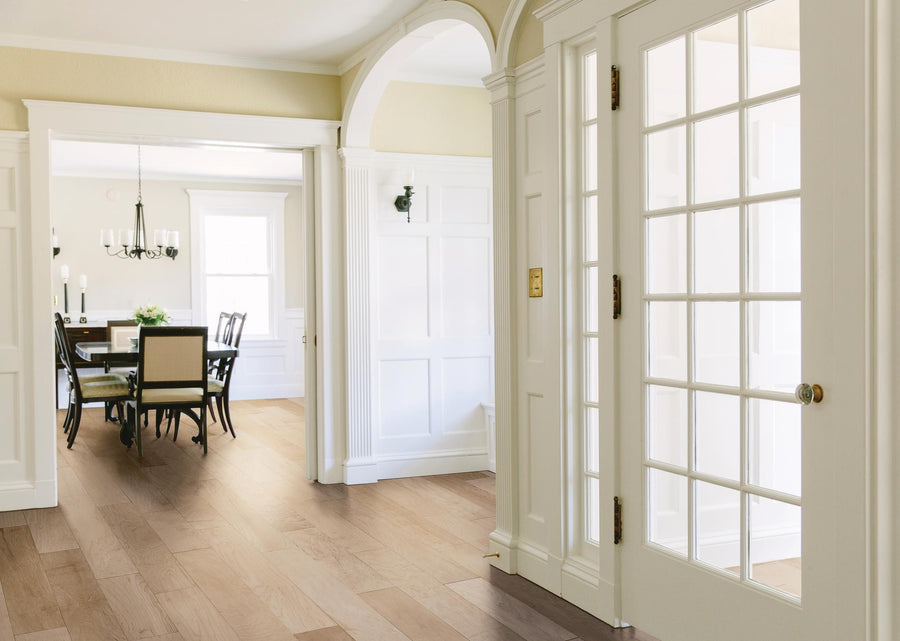 Bright entryway with glass-paneled door opens into a dining room featuring a dark wood table, six chairs, chandelier, and large windows. White paneled walls and light wood floors create an airy, elegant atmosphere.