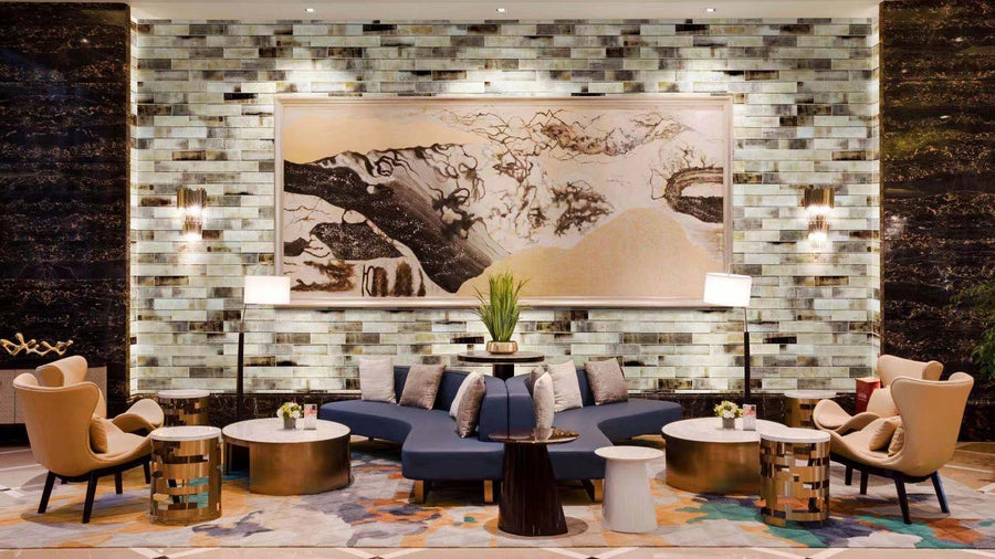 A stylish hotel lobby features modern armchairs, round tables, and a curved blue sofa. A large abstract painting hangs on a tiled wall, with plants and lamps adding to the elegant, contemporary decor.
