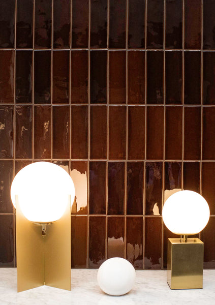 Three spherical lamps, two lit and mounted on gold and brass stands, and one unlit, rest on a white marble surface in front of a glossy, dark brown tiled wall.