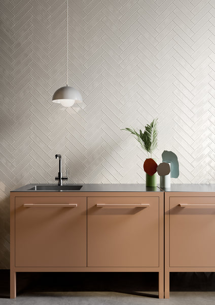 A modern kitchen with light brown cabinets, a sleek black faucet, a white pendant lamp, and minimalist decor including a vase with green leaves on a gray countertop. The backsplash features white herringbone tiles.