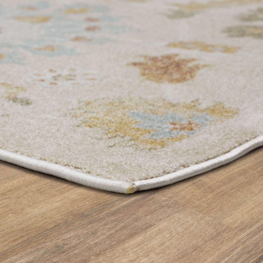 A close-up of the corner of a beige and pastel-patterned area rug on a wooden floor, showing the rug’s soft texture and neatly stitched edge.