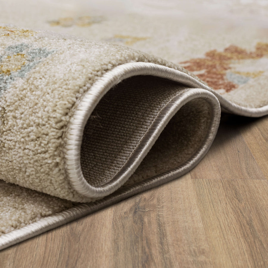 A close-up view of a light-colored rug with a soft texture, partially rolled on a wooden floor, showing the rug’s edge and thickness.
