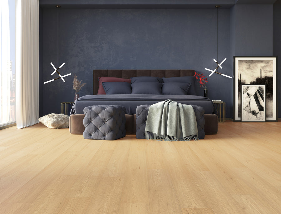 Modern bedroom with dark blue walls, a large bed with navy bedding, two tufted ottomans, a green blanket, abstract framed art on the floor, and light wooden flooring. Natural light comes from tall windows on the left.