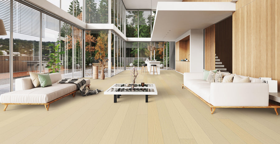 A modern, open-concept living room with large floor-to-ceiling windows, light wood floors, white sofas, a central coffee table, and dining area in the background. Trees and greenery are visible outside.