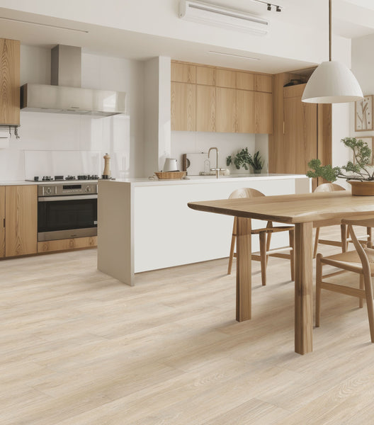 A modern kitchen with light wood cabinets, a white island, built-in oven, and a dining table with chairs. The room features wood flooring, minimalist decor, and natural light from large windows.