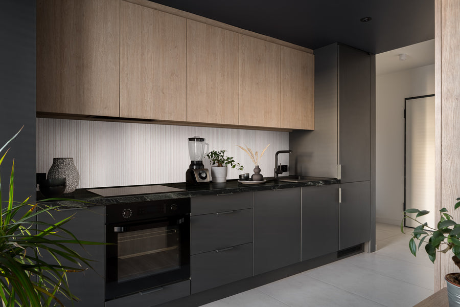 Modern kitchen with dark gray lower cabinets, light wood upper cabinets, built-in oven, blender, plants, and minimalist decor. The space is clean and well-lit with contemporary finishes.