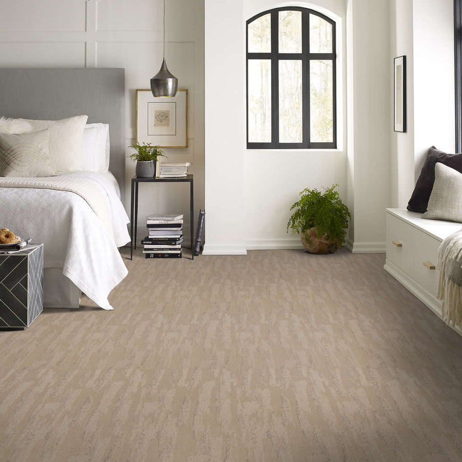 A modern bedroom with a large bed, neutral bedding, a nightstand with decor, a window seat, a potted plant, and beige textured carpet flooring beneath. Light streams in through a tall arched window.