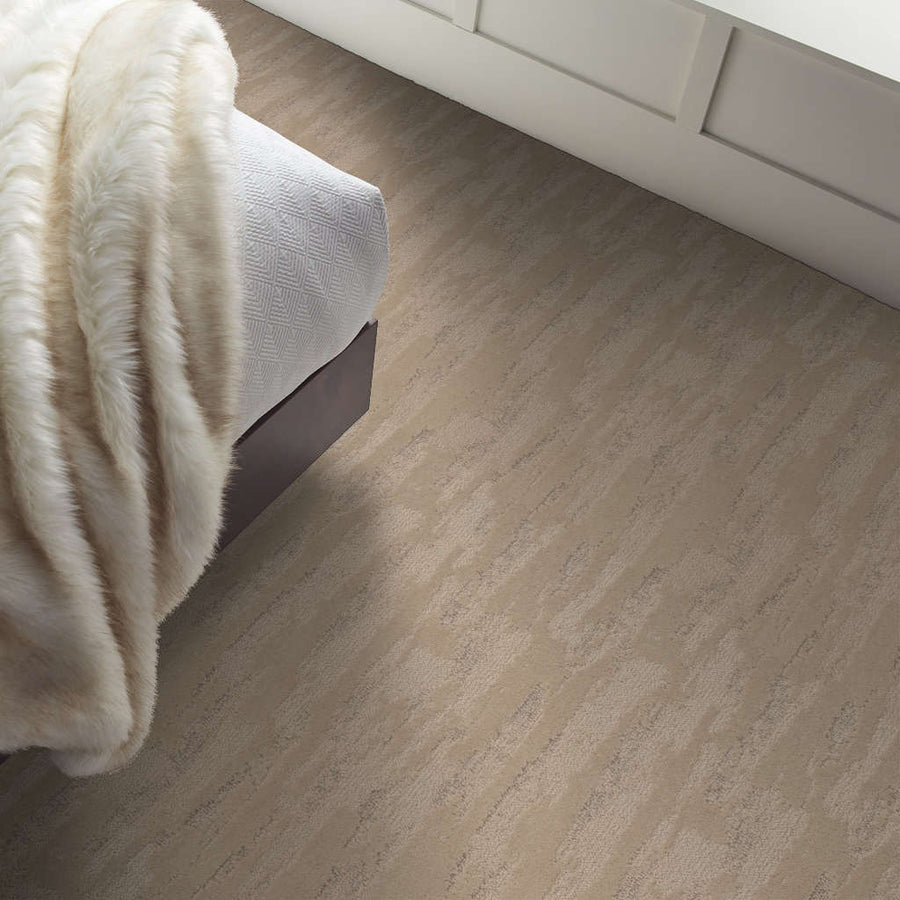 A beige, textured carpet in a bedroom with a bed partially visible, covered with a white blanket and a soft, cream-colored faux fur throw. White paneled walls are seen in the background.
